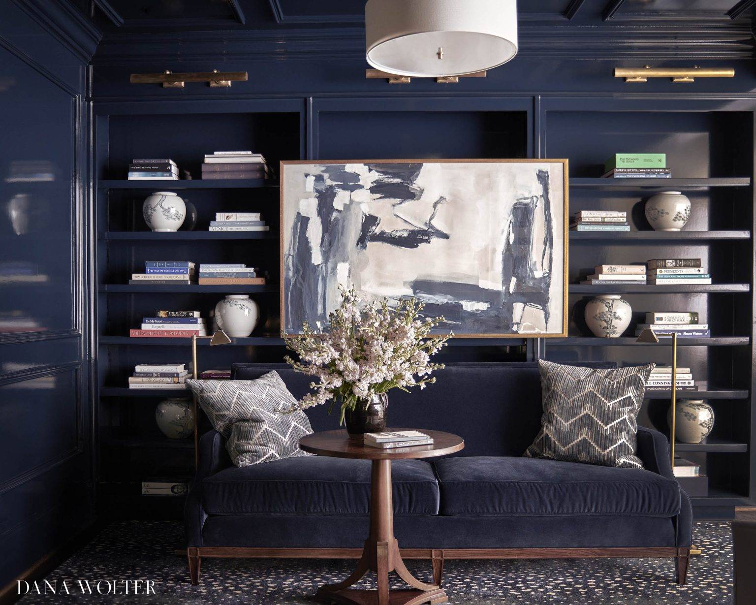 A dark navy living room features a navy velvet sofa with patterned pillows, a round wooden table with a floral arrangement, abstract art, built-in shelves with books, and decorative vases, under a modern white ceiling light.