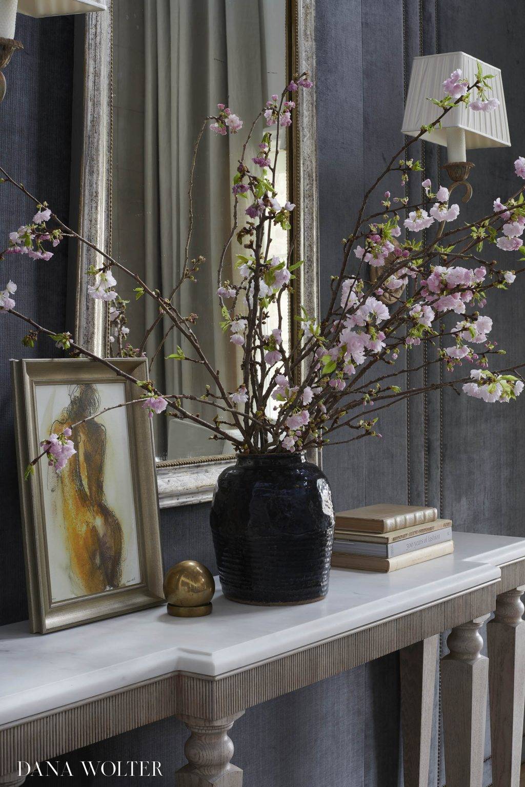 A marble console holds a black vase with pink cherry blossoms, a gold-framed painting of a figure, stacked books, and a gold sphere. A large mirror and two sconces with pleated shades hang on a dark wall behind the console.