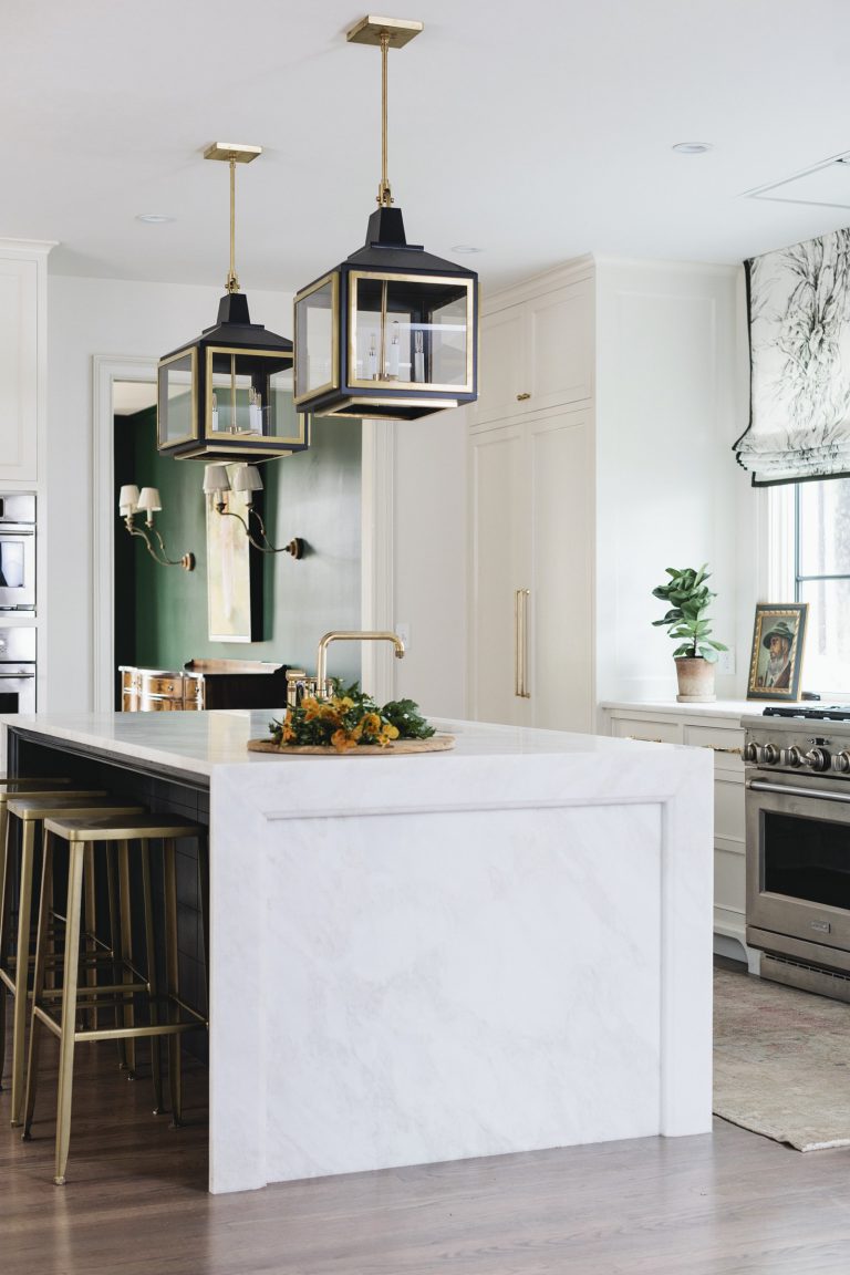 Modern kitchen with a white marble island, brass stools, and two geometric pendant lights. A gold faucet sits on the island with a tray of greenery. White cabinets, stainless steel oven, and a plant on the side counter. Green dining room visible in background.