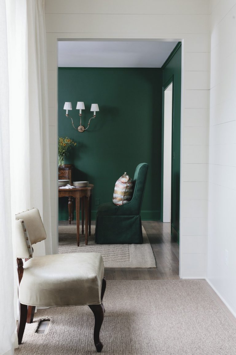A small beige chair sits on a tan carpet in a bright room. Through an open doorway, a green-walled room with a dark green velvet chair, a wooden desk, and a two-light sconce is visible. Curtains hang at left, filtering daylight.