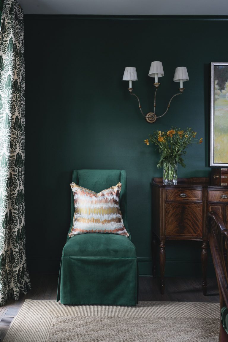 A green velvet chair with a colorful patterned pillow sits against a dark green wall. Above it is a brass wall sconce with two white lampshades. To the right is a wooden sideboard with a vase of yellow flowers. Patterned curtains and a beige rug complete the scene.